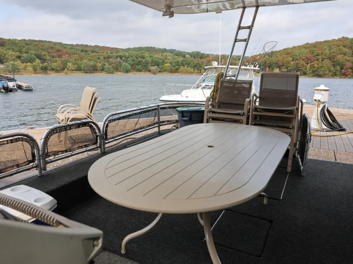 Boat deck with table and chairs, docked near a lake with wooded shoreline.