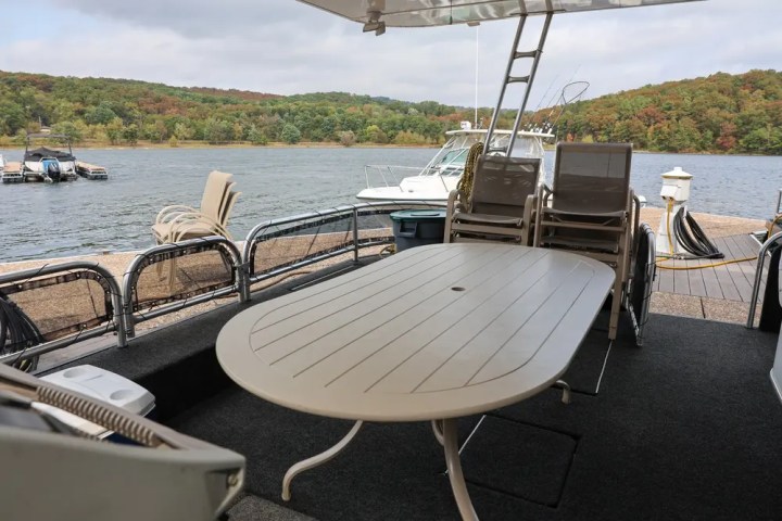 Boat deck with table and chairs, docked near a lake with wooded shoreline.
