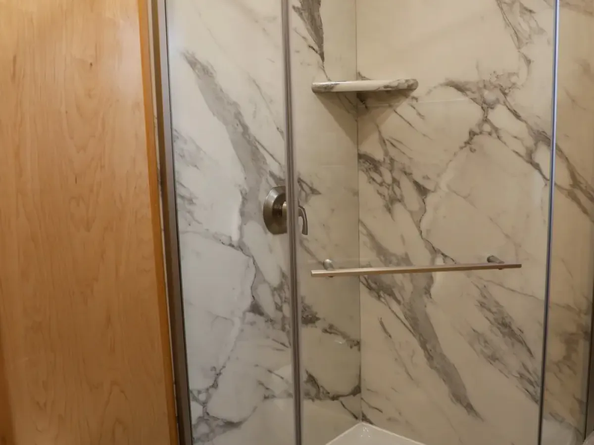 Compact bathroom with corner shower, glass door, marble walls, and adjacent toilet.