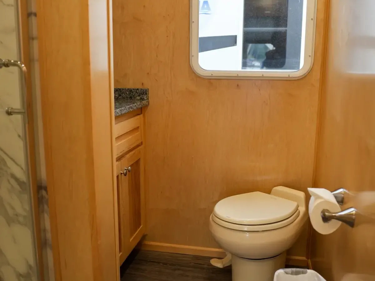 RV bathroom with toilet, sink, shower, window, and trash bin.