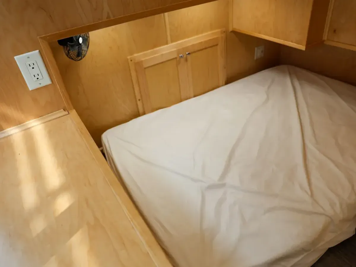 Small wooden bedroom with a bed, shelf, and power outlet on the wall.