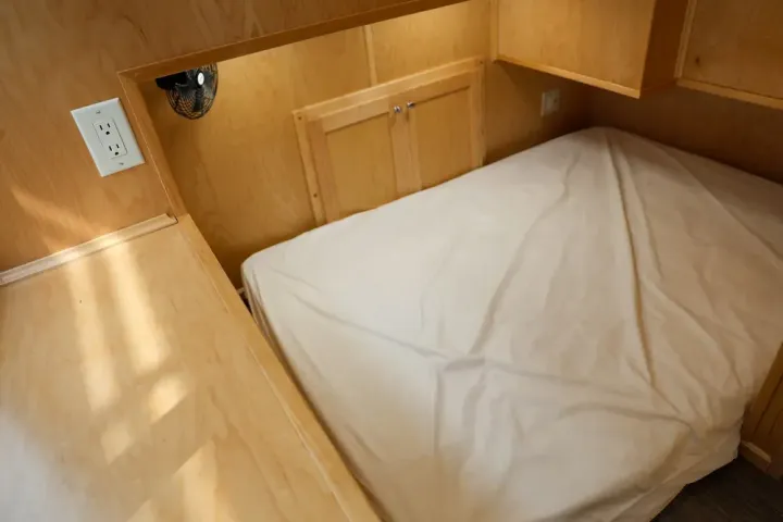 Small wooden bedroom with a bed, shelf, and power outlet on the wall.