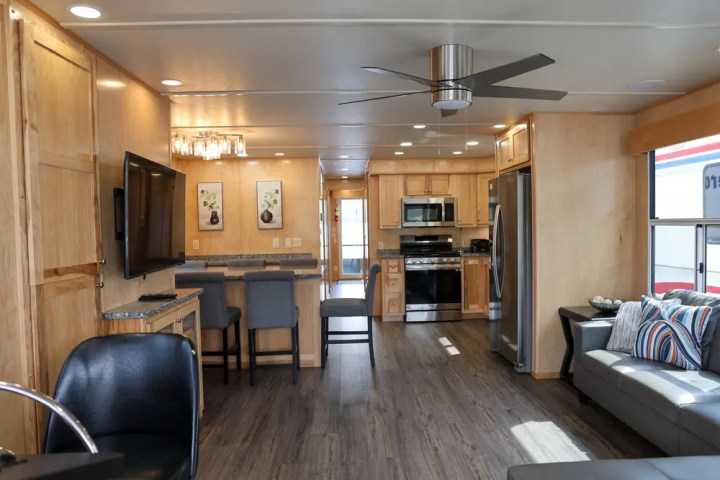 Modern RV interior with wood cabinets, stainless appliances, a sofa, ceiling fan, and mounted TV.