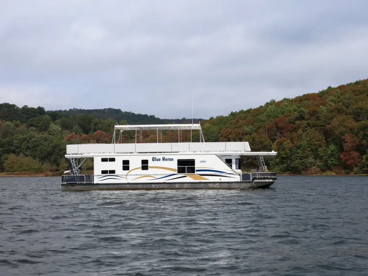 Houseboat named Blue Heron on a lake with forested hills in the background.