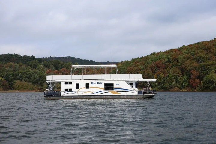 Houseboat named Blue Heron on a lake with forested hills in the background.
