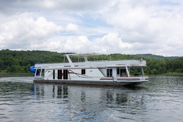 White houseboat on a calm lake with a forested shoreline.