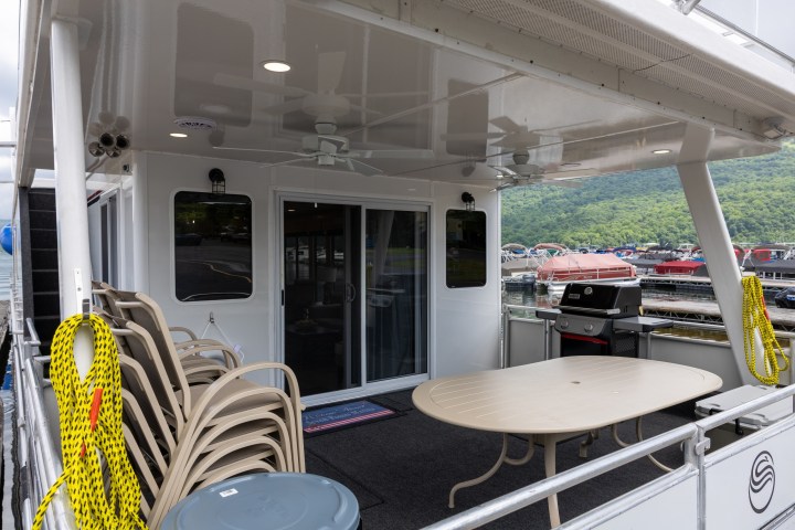 Boat deck with stacked chairs, table, and nearby grill on a lake.