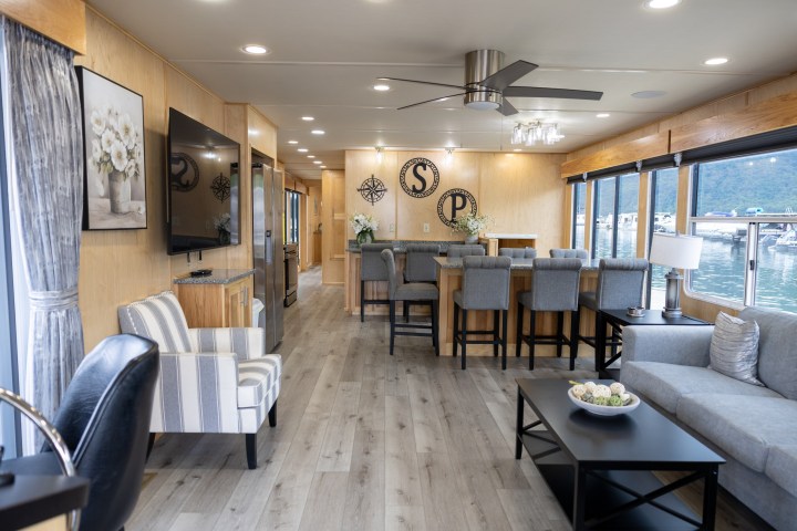 Modern houseboat interior with seating, dining table, and large windows overlooking water.